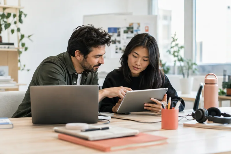 Two software developers reviewing a tablet application alongside a laptop in a bright product studio, focused expressions, coral desk accessory, believable equipment and cables, screens indistinct. AI-generated workplace illustration.