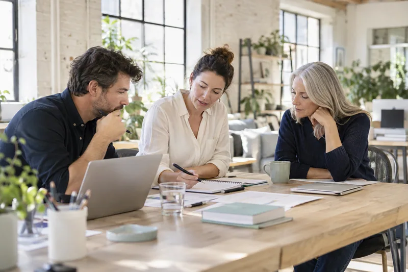 A small owner-led professional services team of three, ages 30 to 60, candidly reviewing tomorrow's work around a light oak desk in a sunlit converted warehouse office. AI-generated workplace illustration.