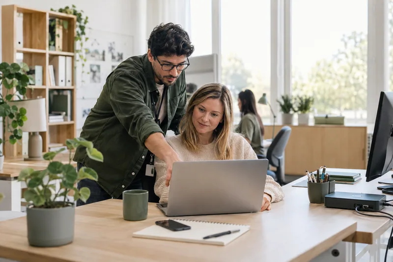 An approachable IT technician helping an employee at their laptop in a modern education administration office, practical equipment and a tidy docking station, muted green details, no server-room fantasy. AI-generated workplace illustration.